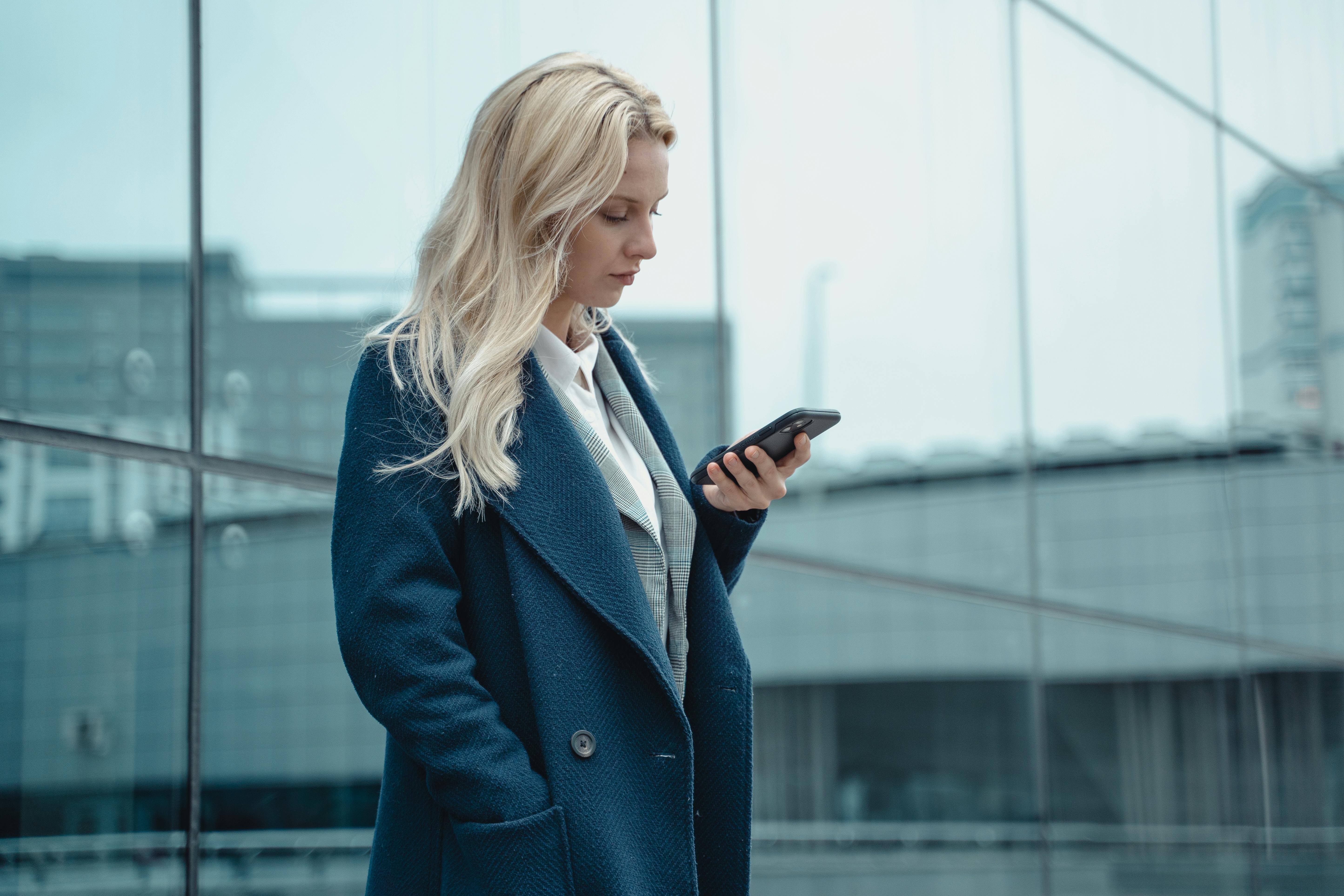 Professional woman in blue coat using smartphone outside office building.